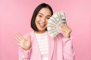 Microcredit, investment and business people concept. Young asian businesswoman, corporate lady showing money, cash dollars, waving hand, pink background photo
