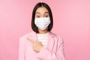 Portrait of asian businesswoman in medical face mask and suit, pointing finger left, showing advertisement, company banner, studio pink background photo