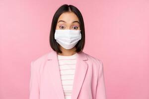 Portrait of asian businesswoman in medical face mask, wearing suit, concept of office work during covid-19 pandemic, standing over pink background photo