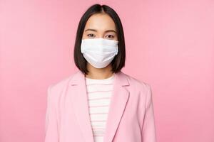 Portrait of asian businesswoman in medical face mask, wearing suit, concept of office work during covid-19 pandemic, standing over pink background photo
