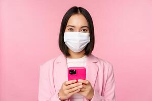 Business people and covid-19 concept. Japanese corporate office lady in suit and medical face mask, using mobile phone and smiling at camera, pink background photo