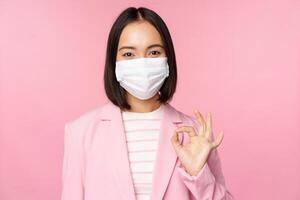 Portrait of asian businesswoman in medical face mask, showing okay sign, wearing suit, work rules during covid-19 pandemic, standing over pink background photo