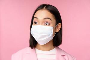 Close-up portrait of asian businesswoman in medical face mask, looking surprised, standing in suit over pink background photo