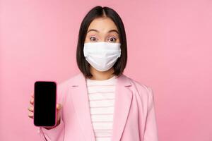 Portrait of smiling korean saleswoman in medical face mask, business suit, showing smartphone screen, standing over pink background photo