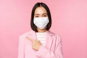 Portrait of asian businesswoman in medical face mask and suit, pointing finger left, showing advertisement, company banner, studio pink background photo