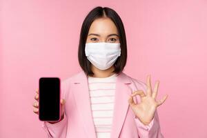 Portrait of asian businesswoman in medical face mask covid-19, showing smartphone screen and okay sign, pink background photo