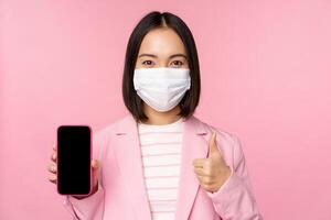 Portrait of asian corporate woman in medical face mask covid-19, showing smartphone screen and thumb up sign, pink background photo