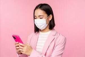 Asian businesswoman in medical face mask using mobile phone. Japenese saleswoman, corporate lady in suit, holding smartphone, standing over pink background photo