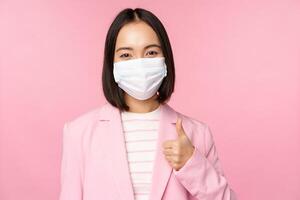 Asian businesswoman in suit and medical face mask, showing thumbs up, recommending wearing personal protective equipment in office during covid-19 pandemic, pink background photo