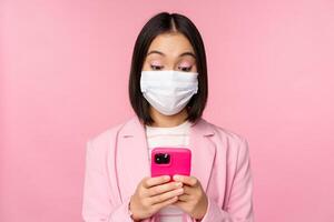 Business people and covid-19 concept. Young asian businesswoman in suit and medical face mask, using smartphone app, standing over pink background photo