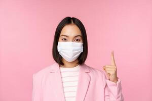 Close up portrait of asian businesswoman in medical face mask and suit, pointing finger up, showing advertisement, top banner, standing over pink background photo