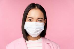 Close up portrait of japanese businesswoman in medical face mask, suit, looking at camera, standing over pink background photo