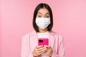 Business people and covid-19 concept. Young asian businesswoman in suit and medical face mask, using smartphone app, standing over pink background photo