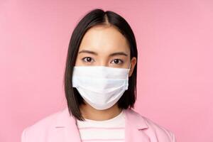 Close up portrait of japanese businesswoman in medical face mask, suit, looking at camera, standing over pink background photo