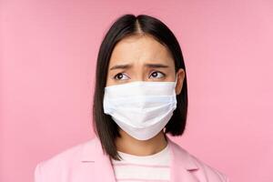 Close up portrait of sad, frowning asian businesswoman in medical face mask, looking left at copy space with disappointed, upset emotion, standing over pink background photo