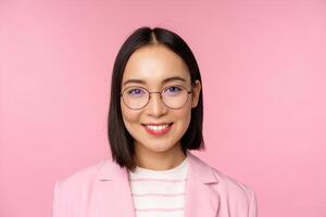 Close up portrait of asian corporate woman, professional businesswoman in glasses, smiling and looking confident at camera, pink background photo