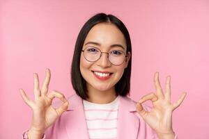 Close up portrait of impressed corporate woman, asian business lady in glasses, showing okay sign, looking amazed at camera, recommending, pink background photo