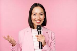 asiático mujer de negocios dando discurso, participación micrófono y sonriente, en pie en traje terminado rosado antecedentes foto