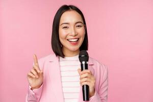 Image of enthusiastic asian businesswoman giving speech, talking with microphone, holding mic, standing in suit against pink studio background photo