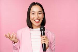 imagen de entusiasta asiático mujer de negocios dando discurso, hablando con micrófono, participación micrófono, en pie en traje en contra rosado estudio antecedentes foto