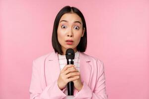 joven asiático vendedora, oficina dama con traje, participación micrófono y mirando conmocionado a cámara, hablando, dando discurso, en pie terminado rosado antecedentes foto