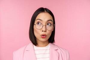 Enthusiastic saleswoman, asian businesswoman in glasses, looking intrigued at upper left corner logo, company advertisement, standing over pink background photo