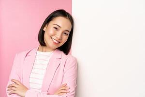 seguro, exitoso japonés oficina dama en traje, cruzar brazos, mirando como profesional a cámara, propensión en blanco pared con anuncio publicitario, vacío Copiar espacio para logo, rosado antecedentes foto