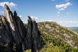 panorámico ver de el montañas en un soleado día. agudo picos en el medio de un verde bosque y pequeño nubes en un claro azul cielo. foto