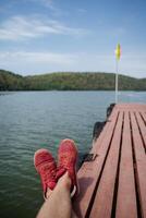 A man rests with his legs folded in red sneakers on the pier. Ahead you can see the yellow flag and green mountains. photo