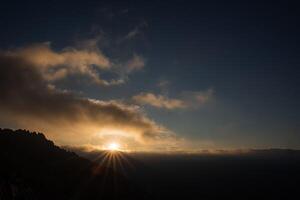 Fascinating panoramic view of the dawn in the mountains. A single large and obese cloud floats across the sky, through which the rays of the sun pass. Shadows from mountains and trees. photo