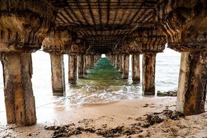 Wooden pillars under the pier. Bright sea water and foam near the shore. Walk by the sea. A shot with a breathtaking perspective photo