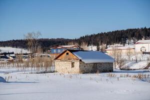 un Roca casa con un de madera cerca en el medio de un cubierto de nieve campo en un soleado día. el techo es de madera y cubierto con nieve. alto calidad foto. allí son varios más grande casas cercano. foto