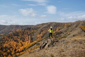 un chico en brillante ropa de deporte camina a lo largo el parte superior de el montaña, mira adelante a el montañas y bosque, claro cielo gastos generales, otoño paisaje foto