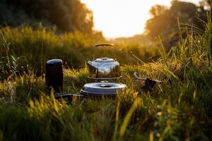 The kettle is on the heater, next to the pot and thermos, all the kitchen utensils for the hike lies in the grass. Sunset illuminates the grass and kettle. Meals in nature, outdoor recreation photo