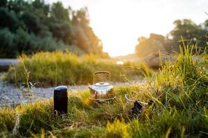 A close shot of a kettle and a thermos by the river bank in the grass. Dinner in nature, meal of a tourist. Hike to nature to the river. Kettle, thermos. photo