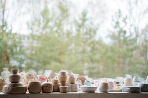 Many ceramics, mainly mugs and vases, stand on a bright windowsill in the workshop. Outside the window there are green trees. photo