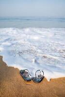 Blue beach slippers on a sandy beach. The wave washes the flip-flops off the shore. Rest by the sea, summer adventures. Surf noise and sea foam photo