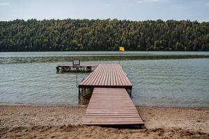 A wooden pier with a yellow flag stands on a sandy beach by the lake. photo