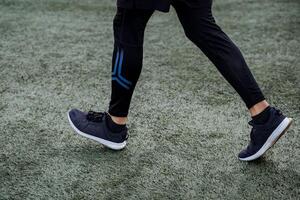 un cerca Disparo de el piernas de un hombre corriendo en un fútbol americano verde campo. Deportes Zapatos y ropa de negro color. al aire libre actividades, Deportes y jugando fútbol. foto