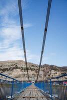 A new and sturdy wooden suspension bridge over a wide river goes to the mountain. A shot with a magnificent perspective, on the shore there are houses. photo