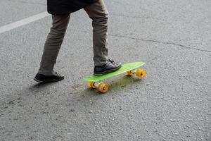 A man in a coat and boots tries to stand on a bright green with yellow wheels pennyboard, skateboard. A hipster learns to skateboard on the asphalt. Activities and new entertainment photo