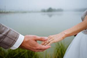 A close shot of the newlyweds, who stretch out their hands with rings to each other. Walk of the groom and the bride near the foggy lake. photo