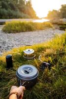 Meal in nature. Pot in the hands of a tourist. Ahead of the river landscape and green forest. Nearby lies the rest of the kitchen camping utensils. Tourist snack, outdoor activities, summer adventure. photo