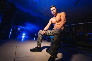 Man rests in gym after having a workout. Strong male with perfect body in the dark background with smoke. Closeup. photo