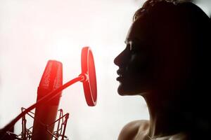 Young sensual silhouette profile with pretty face singing at retro microphone on white background. Sexy woman in front of the mic with red light leaks in studio. Close-up photo