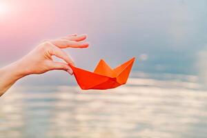mano de un joven mujer participación un papel barco por su dedos encima el río en el verano. naranja papel barco en mujer mano. de cerca foto