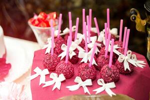 Tasty cake pops on pink sticks decorated with white bows on the table. Wedding buffet with delicious bright cake pops on burgundy napkin. Close-up photo