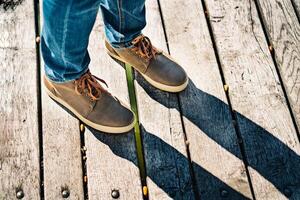 Brown shoes of a traveller on the wooden surface outside. Male's legs in jeans and boots with laces and its shadow on a bridge. Close-up photo