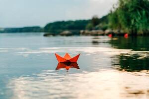 Red paper origami boat floats on the surface of the water and reflects itself on the natural background. Paper ship sailing on blue water surface. photo