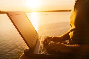 Man works on his laptop in the evening outdoors on the river background. Busy male typing by his hands on a laptop at sunset. Close-up photo